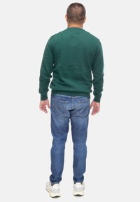 Maglione verde lavorato a maglia, collo a giro, maniche lunghe; abbinato a jeans in denim blu, con finitura testurizzata e cuciture arancioni sulle tasche.