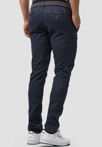 Navy blue chinos with a slim fit, featuring a textured waistband, rear pockets, and subtle stitching details, paired with white sneakers.