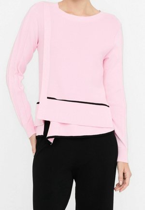 Vrouw draagt een lichtroze lange mouwen gelaagde trui met zwarte afwerkingsdetails, gecombineerd met een zwarte broek.