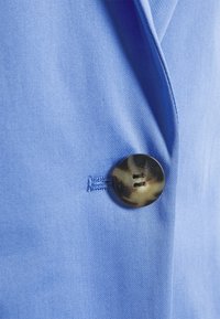 Tissu bleu clair à texture lisse ; présente un grand bouton rond en écaille de tortue avec quatre trous. Bouton cousu sur le côté gauche.