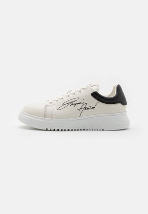 Baskets blanches avec un accent noir au niveau du talon, tige en cuir lisse, bout rond, semelle plate blanche et détail signature sur le côté.