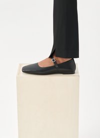 Pantof black din piele cu vârful pătrat, curea la glezne și cataramă argintie. Asortați cu pantaloni negri tapered, stând pe un pedestal din lemn deschis.