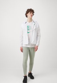 Biely ľahký kabát s kapucňou, zelené tričko s grafikou, světlozelené priliehavé nohavice a čierne tenisky. Jednoduchý, športový dizajn po celom.