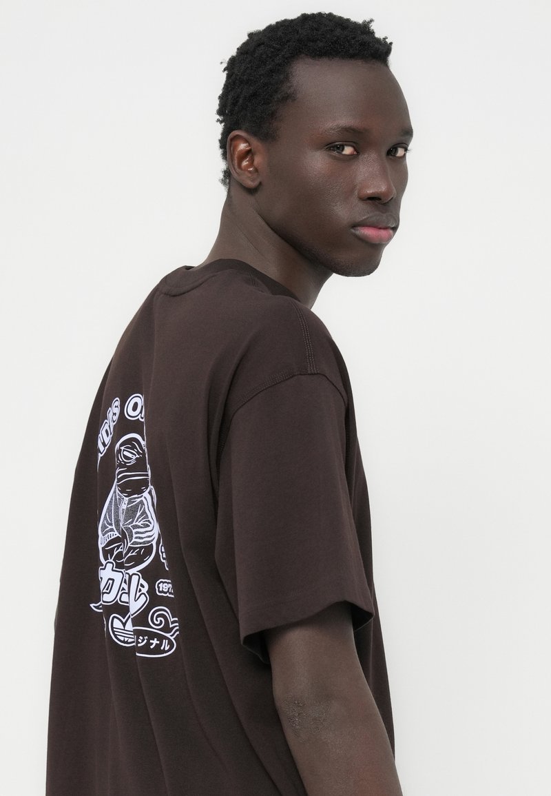 T-shirt marron oversize à manches courtes, avec un imprimé graphique blanc au dos. Texture douce, encolure ronde et coupe décontractée.