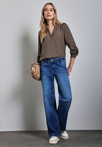 Breda blå jeans med en mjuk textur, kombinerat med en långärmad olivgrön blus och en vävd beige handväska. Vita sneakers fulländar looken.