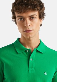 Zelené polo tričko s texturovanou tkaninou, krátkými rukávy a límečkem. Na levé části hrudi má malé vyšívané logo.
