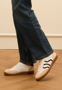 Baskets blanches et beiges avec des accents à rayures noires, semelle en caoutchouc texturée et design à lacets, portées avec un jean bleu foncé sur un sol en bois.