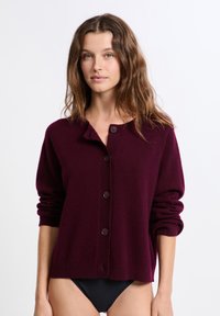 Frau mit welligem braunem Haar, die eine burgunderfarbene, geknöpfte Strickjacke und schwarze Unterteile trägt, vor einem schlichten weißen Hintergrund stehend.