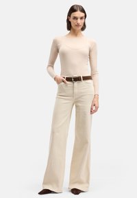 Beige ribgebreide top met lange mouwen, gecombineerd met crèmekleurige wijde broekspijpen. Bruine riem met ronde gesp. Model staat met handen in de zij.