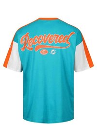 Turkos kortärmad T-shirt med orange detaljer. Texten "Recovered" i fet kursiv på baksidan, med en fotbollslogotyp nedanför.