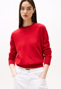 Felpa rossa realizzata in tessuto morbido, con scollatura rotonda e polsini a costine. Presenta un piccolo logo sul petto. Abbinata a jeans bianchi.