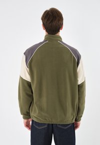 Giacca in pile con colletto alto, caratterizzata da pannelli verde oliva, grigio e crema, e piping bianco lungo le spalle e le maniche.