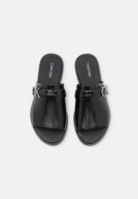 Ett par svarta läder slip-on sandaler från Calvin Klein med öppen tå och en silverfärgad "CK" logotyp på remmarna, visade uppifrån mot en vit bakgrund.