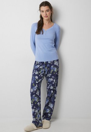 Lichtblauw langslijp henley shirt met drie knopen, gecombineerd met marineblauwe pyjamabroek met bloemenprint in blauw en wit. Crème slippers.