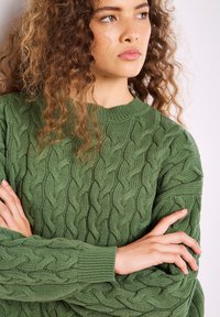 Grön stickad tröja med ett kabelmönster, rund halsringning och ribbade ärmslut med en texturerad yta.