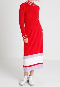 Rotes Strickoberteil mit einem weißen und roten gestreiften Kragen, kombiniert mit einem roten Faltenrock, der horizontale Farbblöcke in Weiß und helllila aufweist.