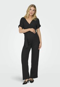 Mono negro con diseño envolvente, mangas cortas y cintura atada. Presenta pantalones de pierna ancha y una tela texturizada. Combinado con zapatos negros.