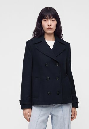Donna con lunghi capelli neri indossa un cappotto doppiopetto blu scuro sopra a una camicia bianca e jeans azzurri su uno sfondo semplice.