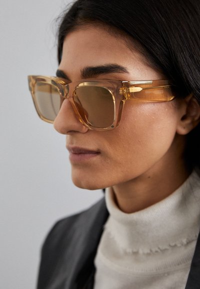 Lunettes de soleil transparentes en ambre avec de grandes lentilles carrées, accents dorés sur la monture et finition brillante. Portées sur un col roulé en tissu léger.