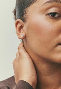 Strieborné náušnice na puzetu v tvare prekrývajúcich sa sŕdc so hladkým povrchom a jemným dizajnom. Modelkina ruka spočíva na krku.