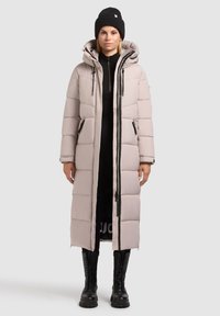 Dlhý puffový kabát v svetlej béžovej farbe, s kapucňou, zipsom v prednej časti a dvoma bočnými vreckami. Skombinovaný s čiernymi bojovými čižmami.