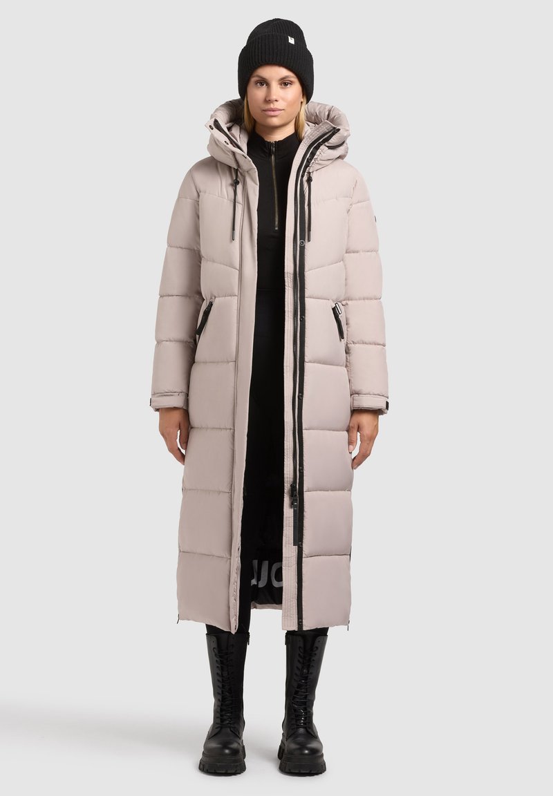 Dlhý puffový kabát v svetlej béžovej farbe, s kapucňou, zipsom v prednej časti a dvoma bočnými vreckami. Skombinovaný s čiernymi bojovými čižmami.