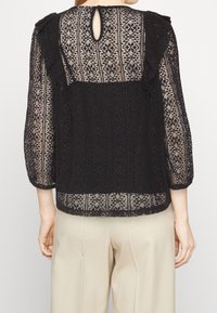 Blusa nera in pizzo caratterizzata da un delicato motivo, spalle arricciate, maniche a tre quarti e design con aperture a chiave sul retro. Abbinata a pantaloni beige.