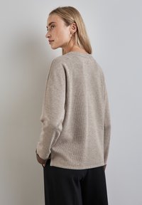 Maglione beige con una texture morbida, scollatura a girocollo e spalle scese. Presenta polsini scuri a contrasto e spacchi laterali per facilitare i movimenti.