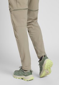 Beige trousers with zippered pockets and a relaxed fit, paired with green hiking shoes featuring a textured sole and yellow accents.
