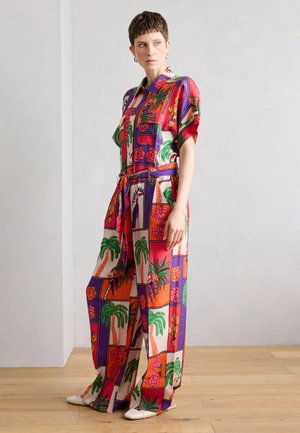 Žena stojaca oblečená v farebne vzorovanom overal s palmami, kvetmi a geometrickými tvarmi, doplnená o biele topánky a náušnice.