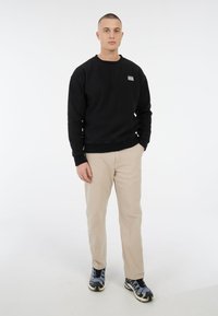 Zwarte sweatshirt met ronde hals, beige broek en gemêleerde zwart-blauwe sneakers. Minimale logo op de linkerkant van de borst. Gladde textuur.