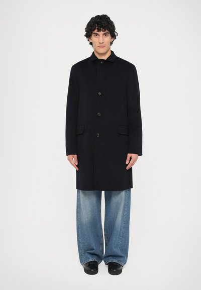 Homme debout, portant un manteau noir boutonné, un jean bleu ample et des chaussures noires, sur un fond blanc uni.