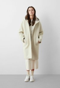 Cappotto lungo beige chiaro con cappuccio, tessuto strutturato, ampie tasche laterali e bottoni in legno, indossato sopra un vestito bianco e sneakers.