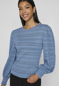 Světle modrá blůza s texturovanými pruhy, kulatým výstřihem, dlouhými nabíranými rukávy a elastickými manžetami. Tkanina má otevřené pletené vzory.