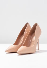 Escarpins en cuir verni nude avec des orteils pointus, des talons fins en stiletto, et une texture lisse, comportant un détail de doublure intérieure subtile.