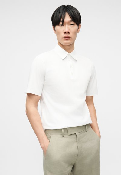 Νεαρός άνδρας με μαύρα μαλλιά που φοράει λευκό πόλο με κοντά μανίκια, τα οποία έχει βάλει μέσα σε ανοιχτό μπεζ παντελόνι, με τα χέρια στις τσέπες.