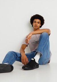 Μαύρα παπούτσια, τζιν ελαφρού πλυσίματος, και γκρι κοντομάνικο μπλουζάκι με σχέδιο. Το άτομο κάθεται χαλαρά με το ένα πόδι λυγισμένο, τα χέρια resting στο γόνατο.