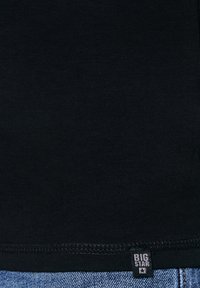 Mustast kangast õmmeldud serv sinise denimi kohal, millel on väike must silt, millel on kiri "BIG STAR" ja tähe sümbol.