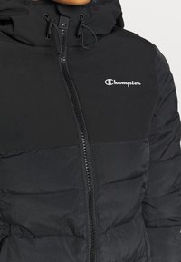 Black puffer jacket with a zip front, featuring a soft texture, a hood, and a small white Champion logo on the chest.