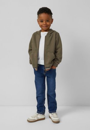 Olijfgroen ritshoodie, wit t-shirt, blauwe denim jeans en witte sneakers met lichtbruine zolen. Model staat met de handen in de zakken.