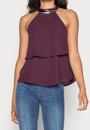 Haut sans manches violet à col halter avec tissu superposé et encolure ornée de sequins, porté avec un jean en denim bleu sur le torse d'une femme.