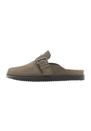 Hnedý semišový nazúvací šľapák s nastaviteľným remienkom a kovovou prackou, čiernou ryhovanou podrážkou a korkovou stielkou viditeľnou pri päte.