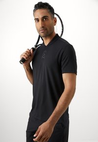 Polo preto com colarinho curto, apresentando um detalhe sutil do logo. O tecido parece suave e leve, ideal para vestuário atlético.
