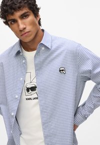 Kockovaná modro-biela košeľa s malou čiernou grafickou nášivkou. Nosená cez biely tričko so skicou osoby.