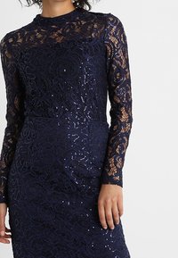 Marinblå spetsklänning med långa ärmar, som har intrikata blommönster och paljetter för textur. Hög halsringning och figurnära design.