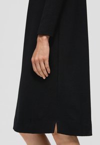 Schwarzes, strukturiertes Kleid mit knielangem Schnitt, langen Ärmeln und einem Seitenschlitz, mit einer Hand, die auf dem Stoff ruht.