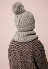 Grijze gebreide beanie met een pluizige pompon en bijpassende geribbelde sjaal, gedragen over een bruine houndstooth-gepatenteerde jas. Gegevens van de gestructureerde stof zijn zichtbaar.