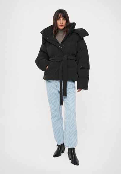 Kvinde iført en sort bælte-buffetjakke, grå turtleneck, lys blå mønstrede jeans og sorte ankelstøvler, stående mod en hvid baggrund.