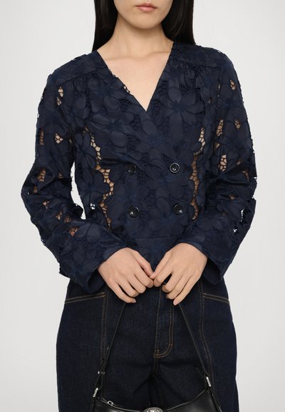 Navy blue, lace blouse with floral patterns, double-breasted design, v-neckline, and long sleeves, paired with dark denim pants and a black handbag.