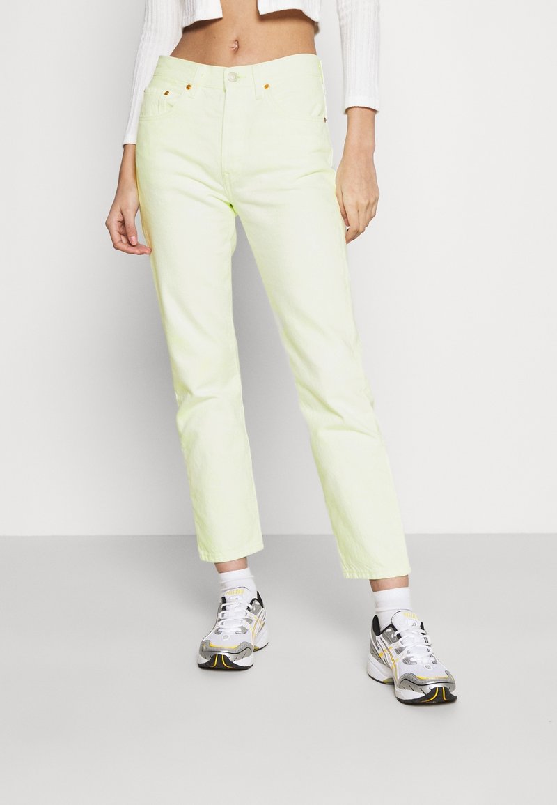 Personne portant un jean droit jaune clair, des baskets blanches avec des détails jaunes et noirs, et un haut court blanc à manches longues.